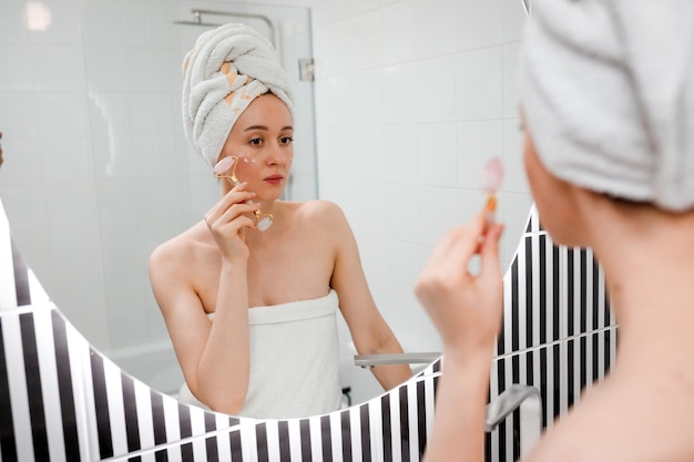 Mooie vrouw met perfecte huid die een witte handdoek draagt na het douchen die gezichtsmassage maakt met behulp van een jade gezichtsroller met natuurlijke kwartssteen in de badkamer Natuurlijke cosmetica concept rimpelvereffening