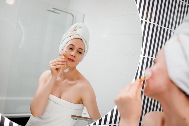 Mooie vrouw met perfecte huid die een witte handdoek draagt na het douchen die gezichtsmassage maakt met behulp van een jade gezichtsroller met natuurlijke kwartssteen in de badkamer Natuurlijke cosmetica concept rimpelvereffening