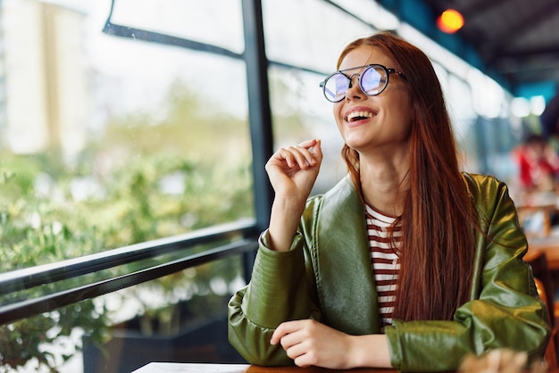Foto mooie vrouw met rood haar en een bril zit in de stad in een café en kijkt uit het raam glimlach met tanden vrouwelijke freelance blogger