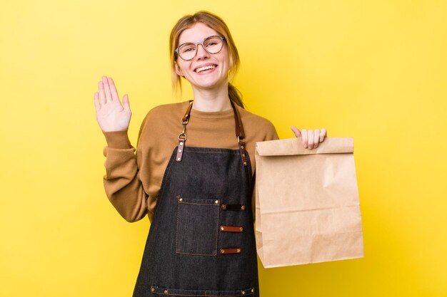 Mooie vrouw met rood hoofd die lacht en vrolijk zwaait met de hand die je verwelkomt en je het bezorgconcept begroet