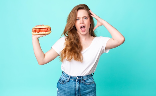 Mooie vrouw met rood hoofd die met handen in de lucht schreeuwt en een hotdog vasthoudt