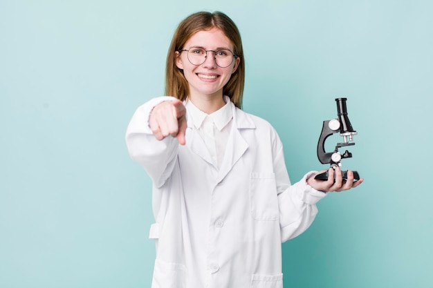 Mooie vrouw met rood hoofd die naar de camera wijst en je wetenschapper kiest met een microscoopconcept