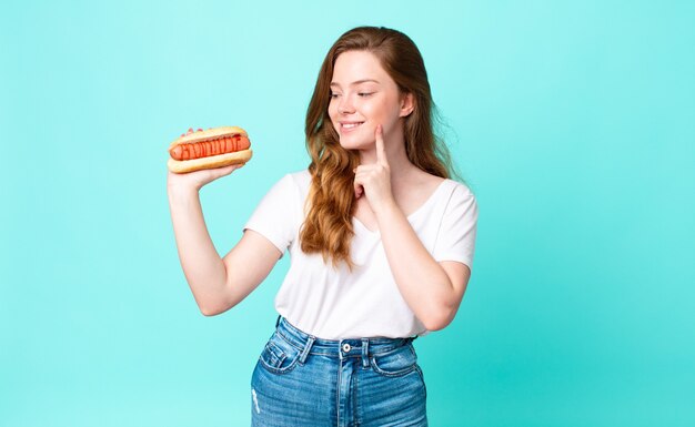 Mooie vrouw met rood hoofd die vrolijk lacht en dagdroomt of twijfelt en een hotdog vasthoudt