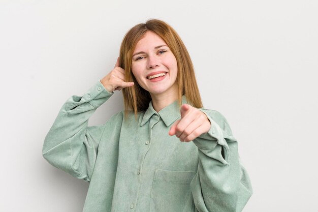 Mooie vrouw met rood hoofd die vrolijk lacht en naar de camera wijst terwijl ze belt, gebaar later aan de telefoon