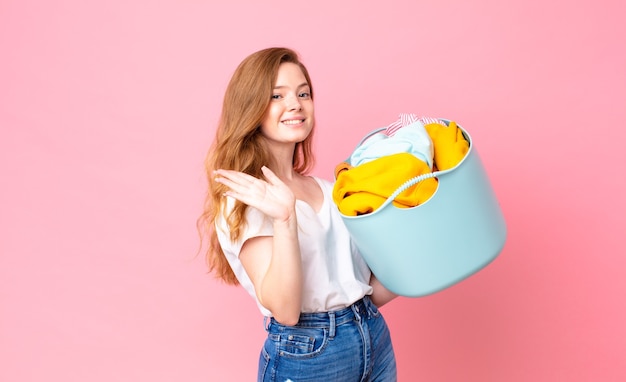 Mooie vrouw met rood hoofd die vrolijk lacht, met de hand zwaait, je verwelkomt en begroet en een wasmand met kleren vasthoudt