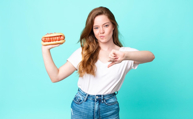 Mooie vrouw met rood hoofd die zich boos voelt, duimen naar beneden laat zien en een hotdog vasthoudt