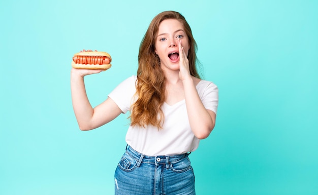 Mooie vrouw met rood hoofd die zich gelukkig voelt, een grote schreeuw geeft met de handen naast de mond en een hotdog vasthoudt