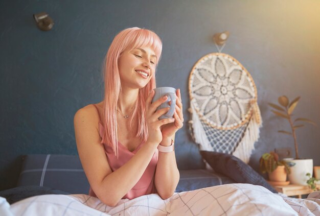 Mooie vrouw met roze haar houdt mok zittend op comfortabel bed met zachte deken