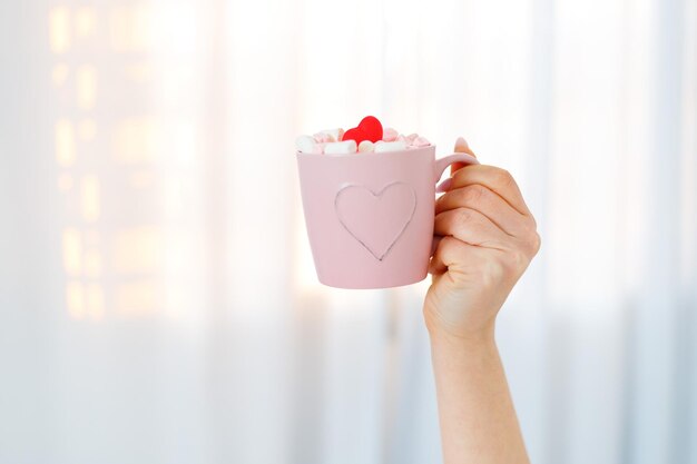 Mooie vrouw met roze kopje cacao marshmallows en hartvormige snoep op bed thuis op Valentijnsdag.