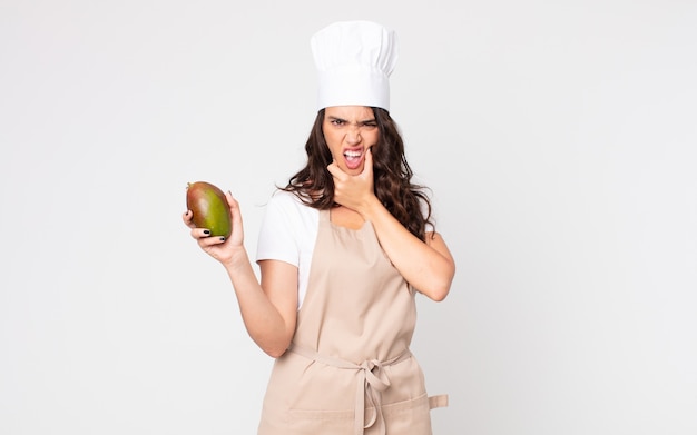 Mooie vrouw met wijd open mond en ogen en hand op de kin die een schort draagt en een mango vasthoudt