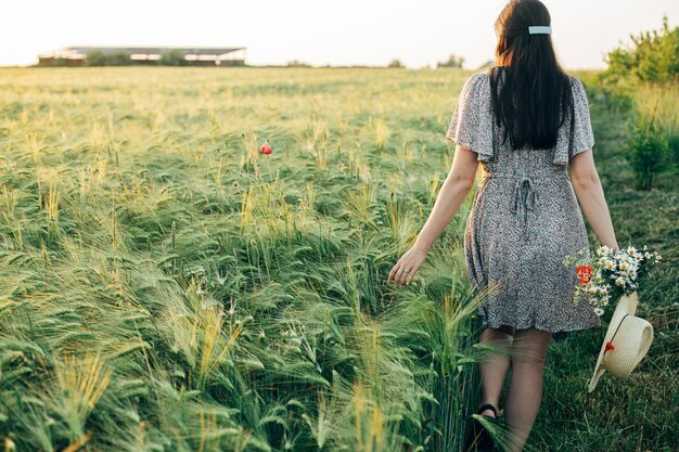 Mooie vrouw met wilde bloemen en strohoed wandelen in gerstveld bij zonsonderganglicht Stijlvolle vrouw ontspannen in de avondzomer op het platteland en bloemen verzamelen Sfeervol rustig moment