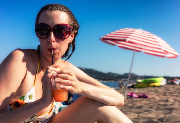 Mooie vrouw met zonnebril in badpak alcoholische drank drinken op zandstrand in resort