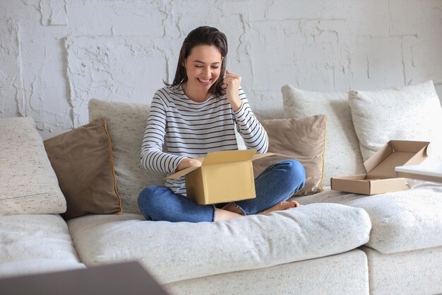 Mooie vrouw pakt pakket uit en drukt vreugde uit terwijl ze thuis op de bank zit