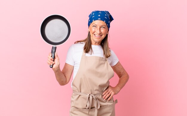 Mooie vrouw van middelbare leeftijd die gelukkig lacht met een hand op de heup en zelfverzekerd en een pan vasthoudt. chef-kok concept