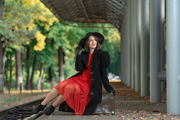 Mooie vrouw van middelbare leeftijd met retro koffer op het treinstation wachtend op trein Vrouw in rode jurk en hoed herfst