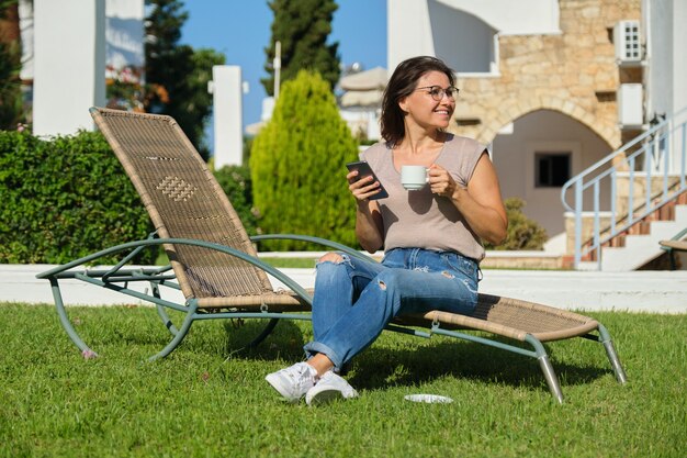 Mooie vrouw van middelbare leeftijd rusten zittend op zonnebank op groen gazon, smartphone lezen via internet, genieten van kopje koffie ontspannen op zonnige zomerdag. Vakanties, levensstijl, vrije tijd van leeftijd mensen