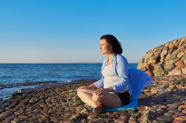 Mooie vrouw van middelbare leeftijd zittend in lotuspositie mediteren aan de kust bij zonsondergang