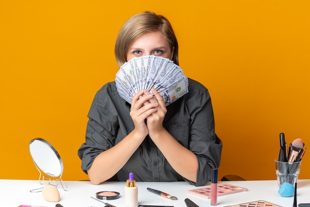 mooie vrouw zit aan tafel met make-up tools bedekt gezicht met contant geld