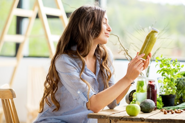 Mooie vrouw zitten met drankjes en gezond groen voedsel thuis. Veganistisch maaltijd- en detoxconcept