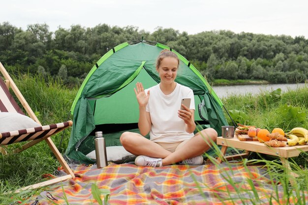 Mooie vrouw zittend in de buurt van tent en rivier actieve levensstijl reizen tijd doorbrengen in de buurt van rivier met smartphone met videogesprek zwaaiend hallo of tot ziens groet