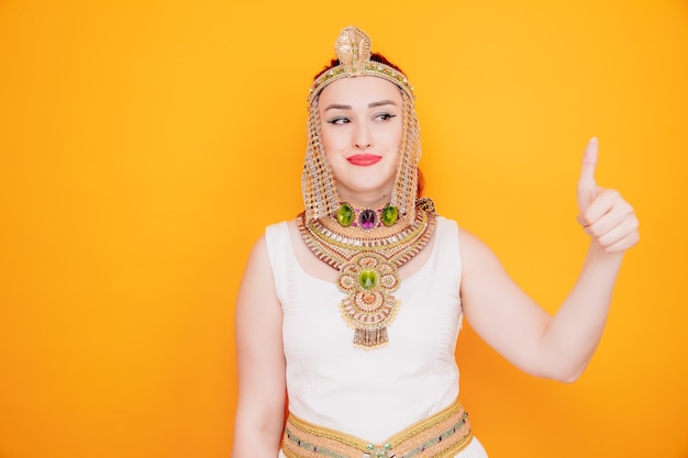 Mooie vrouw zoals cleopatra in oud egyptisch kostuum die opzij kijkt glimlachend zelfverzekerd met duim omhoog op sinaasappel