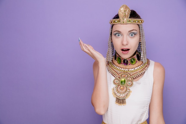 Mooie vrouw zoals cleopatra in oud egyptisch kostuum verbaasd en verrast met opgeheven arm op paars