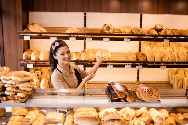 Mooie vrouwelijke arbeider die banketbakkersproducten en brood op planken toont