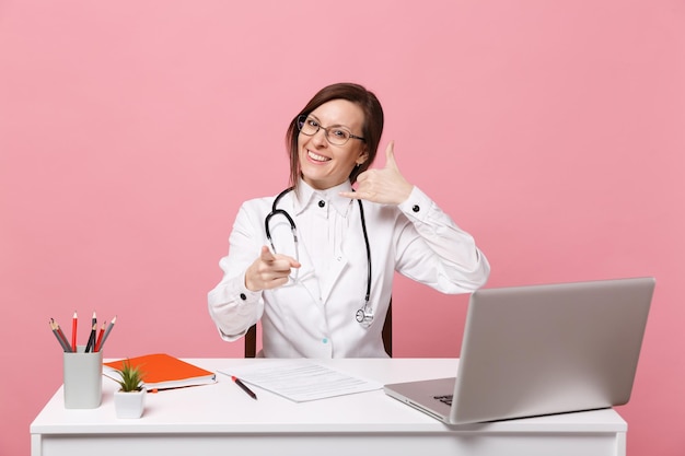 Mooie vrouwelijke arts zit aan bureau werkt op computer met medisch document in ziekenhuis geïsoleerd op pastel roze muur achtergrond. vrouw in medische toga glazen stethoscoop. gezondheidszorg geneeskunde concept