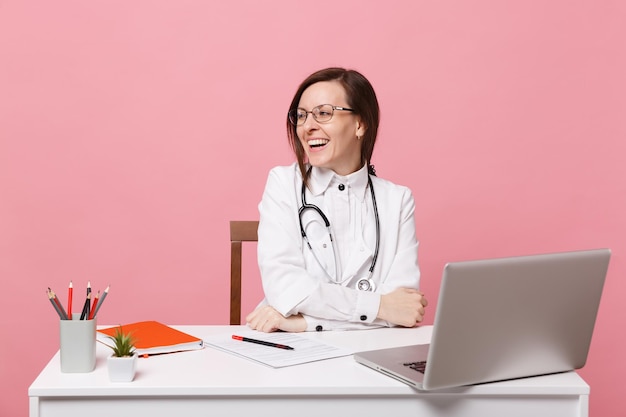 Mooie vrouwelijke arts zit aan bureau werkt op computer met medisch document in ziekenhuis geïsoleerd op pastel roze muur achtergrond. Vrouw in medische toga glazen stethoscoop. Gezondheidszorg geneeskunde concept