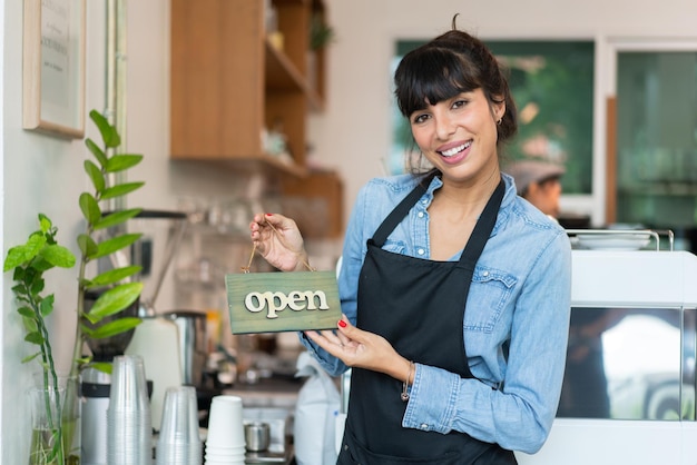 Mooie vrouwelijke barista-eigenaar in schort glimlachend met open bord welkom bij klant in coffeeshop