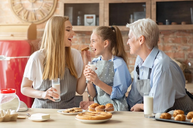 Mooie vrouwelijke familie die gebak met melk eet in de keuken, moeder die dochter voedt, grootmoeder lacht