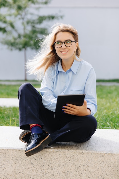 Mooie vrouwelijke hipster in brillen, blauw shirt en zwarte jeans met behulp van digitale tablet op straat. Concept van mensen, technologie en stedelijke levensstijl.