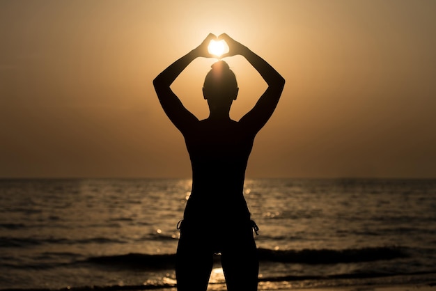 Mooie vrouwelijke Model genieten van zonsondergang en het maken van hart teken op zon aan zee kalm water weerspiegelt silhouet van vrouw zon gaat onder Horizon