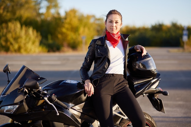 mooie vrouwelijke piloot zittend op zwarte motobike, draagt leren jas, zwarte broek, wit overhemd en heeft rode bandana op nek, stopt onderweg om uit te rusten