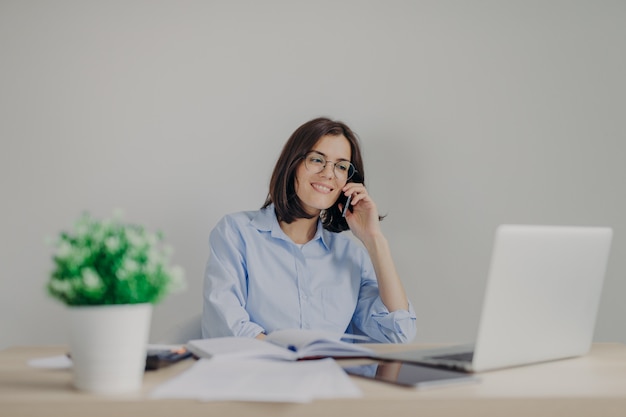 Mooie vrouwelijke recruiter doet jobaanbieding aan somene via mobiele telefoon, controleert curriculum vitae op laptopcomputer