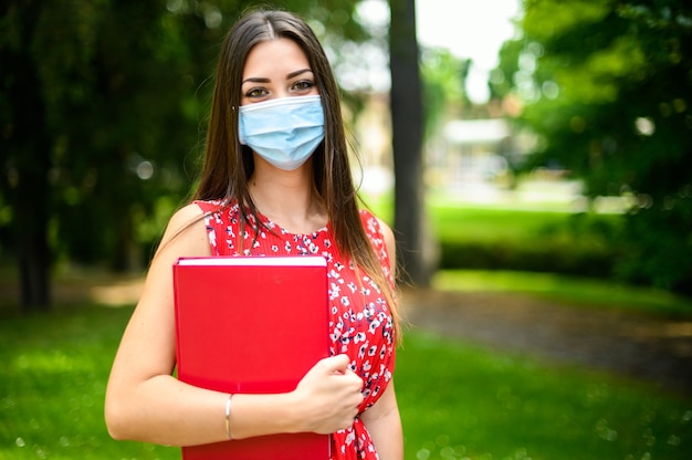 Mooie vrouwelijke student die een boek buiten houdt en een masker draagt, coronavirusconcept