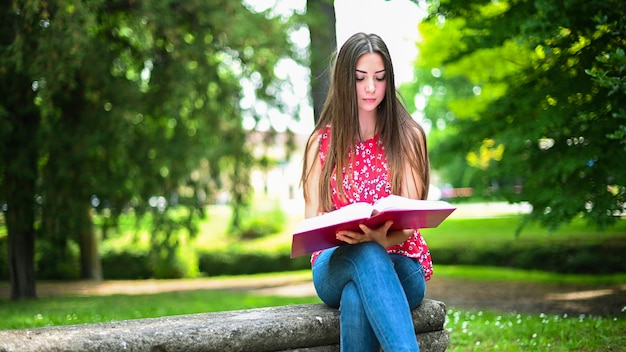 Mooie vrouwelijke student die een boek op een bank in een park leest