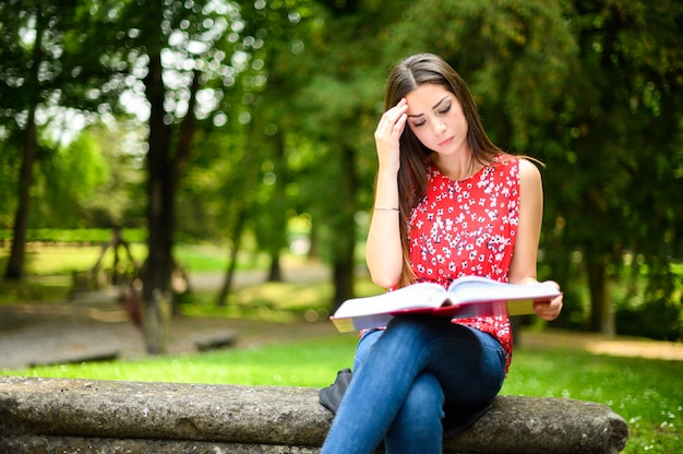 Mooie vrouwelijke student die een boek op een bank in een park leest