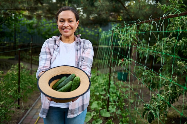 Mooie vrouwelijke tuinman met een brede glimlach die een strohoed vasthoudt met vers geplukte rijpe komkommers op een biologische boerderij