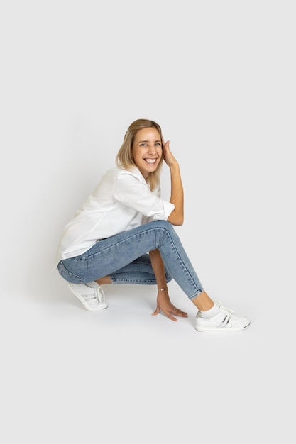 Mooie vrouwelijke volwassene met grote glimlach op gezicht camera kijken en poseren in semisquat op witte achtergrond in witte blouse spijkerbroek en sneakers Positieve emoties tonen