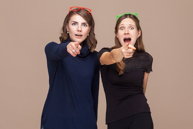 Mooie vrouwen die bij elkaar staan en met de vingers naar de camera en de schok wijzen. Studio opname, lichtbruine achtergrond