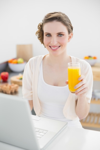 Mooie vrouwenzitting voor haar laptop in haar keuken terwijl het houden van een glas jus d&#39;orange