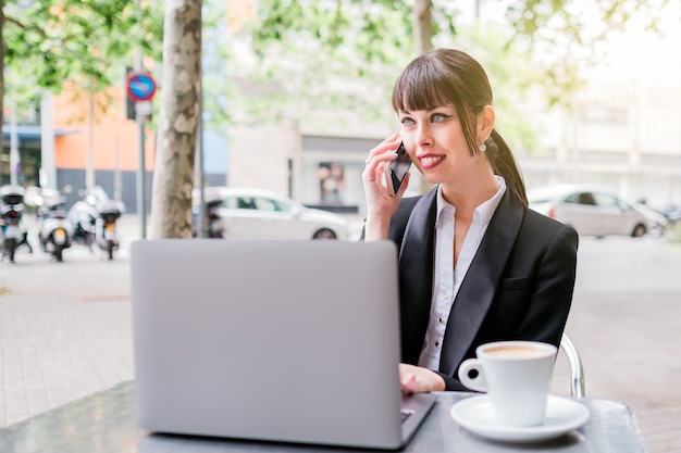 mooie zakenvrouw die een laptop gebruikt die buiten in de coffeeshop zit en smartphone gebruikt