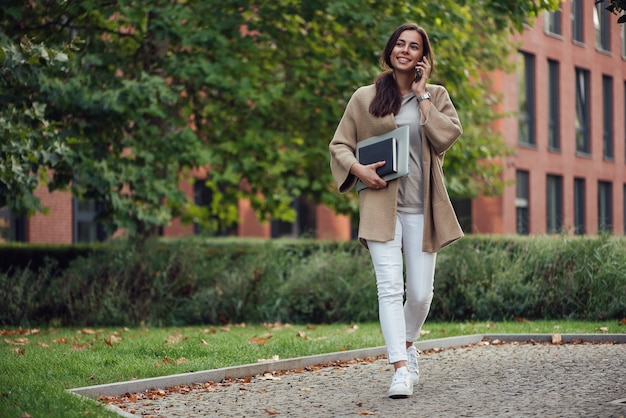 Mooie zakenvrouw in casual outfit met een laptop en notebook buiten spreekt op smartphone