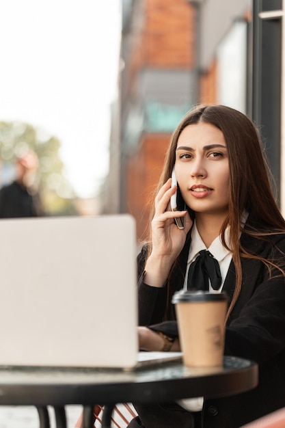 Mooie zakenvrouw in een mode zwart pak zit in een café praten aan de telefoon werken op een laptop en koffie drinken in de stad Jonge professionele hr meisje op straat