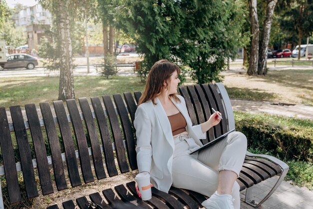 Mooie zakenvrouw in een wit pak zit in een stadspark, drinkt koffie en werkt op een digitale tablet