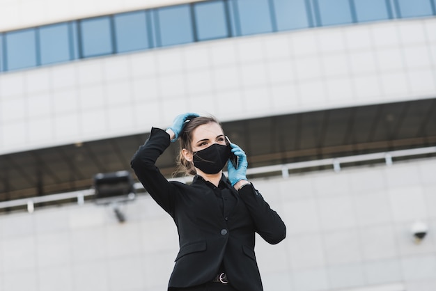 Mooie zakenvrouw in een zwart pak in een zwart medisch masker en handschoenen in de stad in quarantaine en isolatie. pandemie covid-19. afbeelding met selectieve aandacht