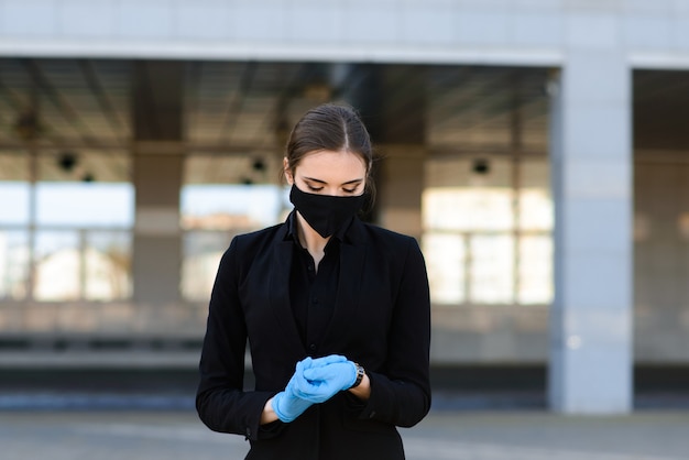 Mooie zakenvrouw in een zwart pak in een zwart medisch masker en handschoenen in de stad in quarantaine en isolatie. Pandemie COVID-19. Afbeelding met selectieve aandacht