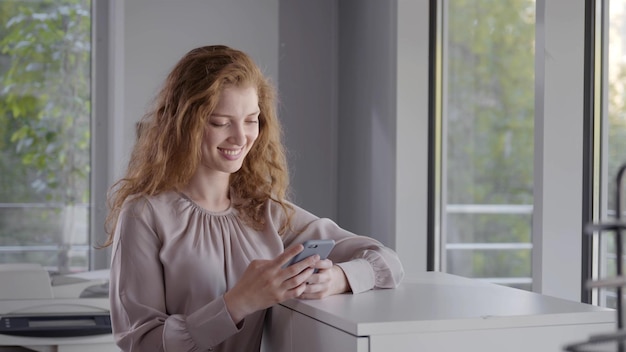 Mooie zakenvrouw met behulp van mobiele telefoon in modern kantoor in de buurt van venster