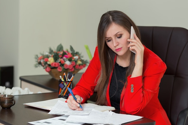 Mooie zakenvrouw praten over de telefoon en schrijven op papier in kantoor.